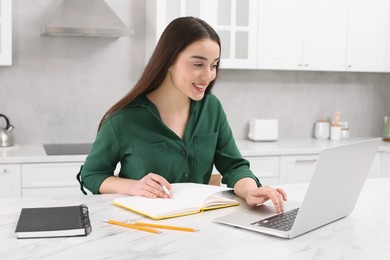 Photo of Home workplace. Happy woman with pen and notebook working on laptop at marble desk in kitchen