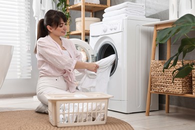 Happy young housewife putting laundry into washing machine at home