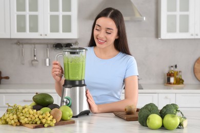 Beautiful young woman preparing tasty smoothie at white table in kitchen