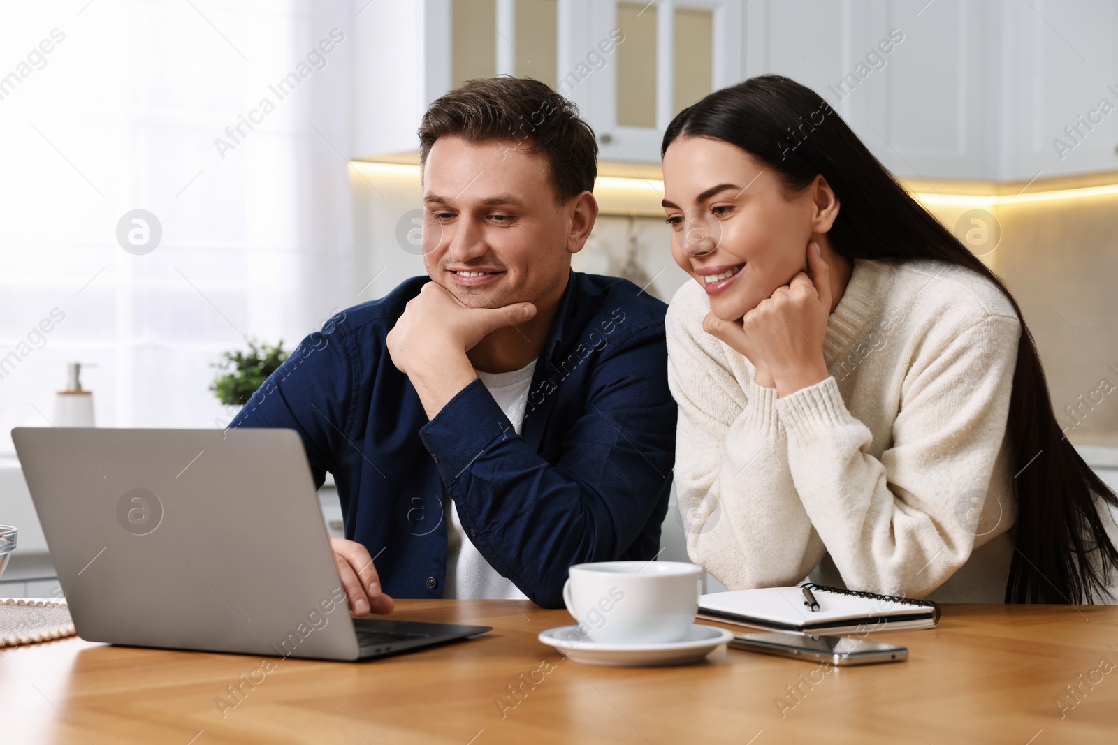 Photo of Happy couple using laptop together at wooden table in kitchen