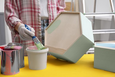 Photo of Woman dipping brush into bucket of green paint at yellow wooden table indoors, closeup
