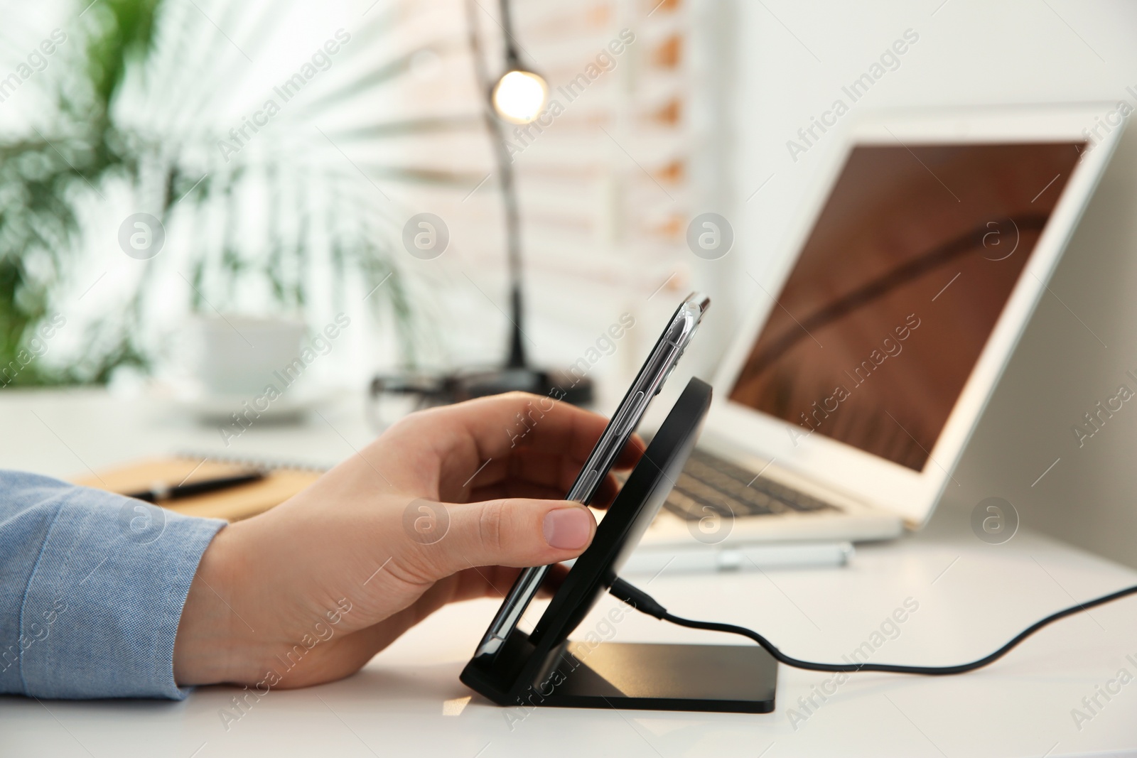 Photo of Man putting mobile phone onto wireless charger at white table, closeup. Modern workplace accessory