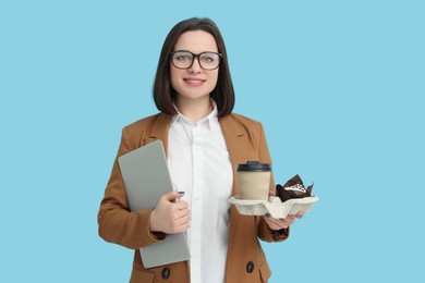 Happy young intern holding laptop, takeaway cup with hot drink and muffin on light blue background