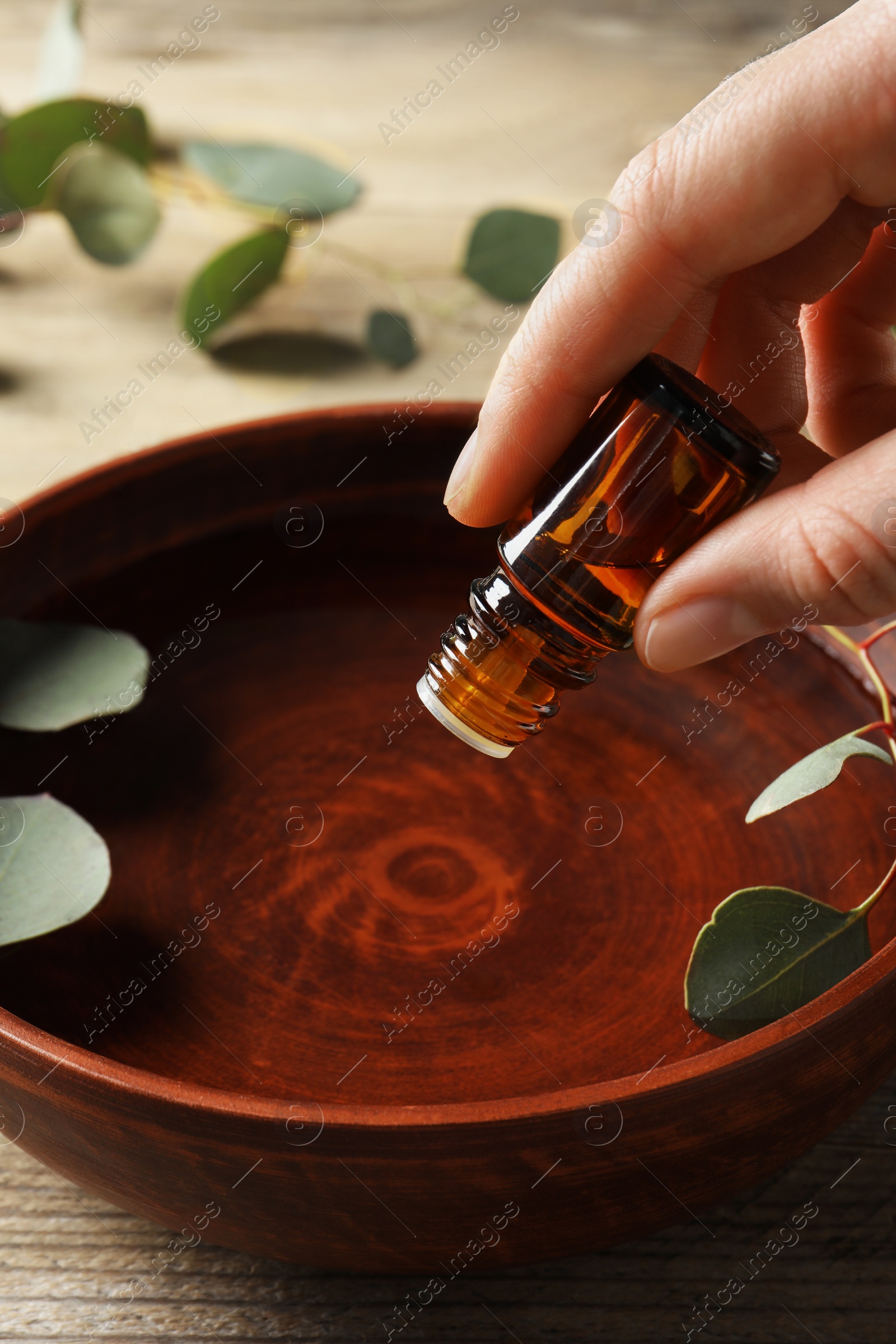 Photo of Woman dripping eucalyptus essential oil from bottle into bowl at wooden table, closeup