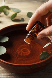 Photo of Woman dripping eucalyptus essential oil from bottle into bowl at wooden table, closeup