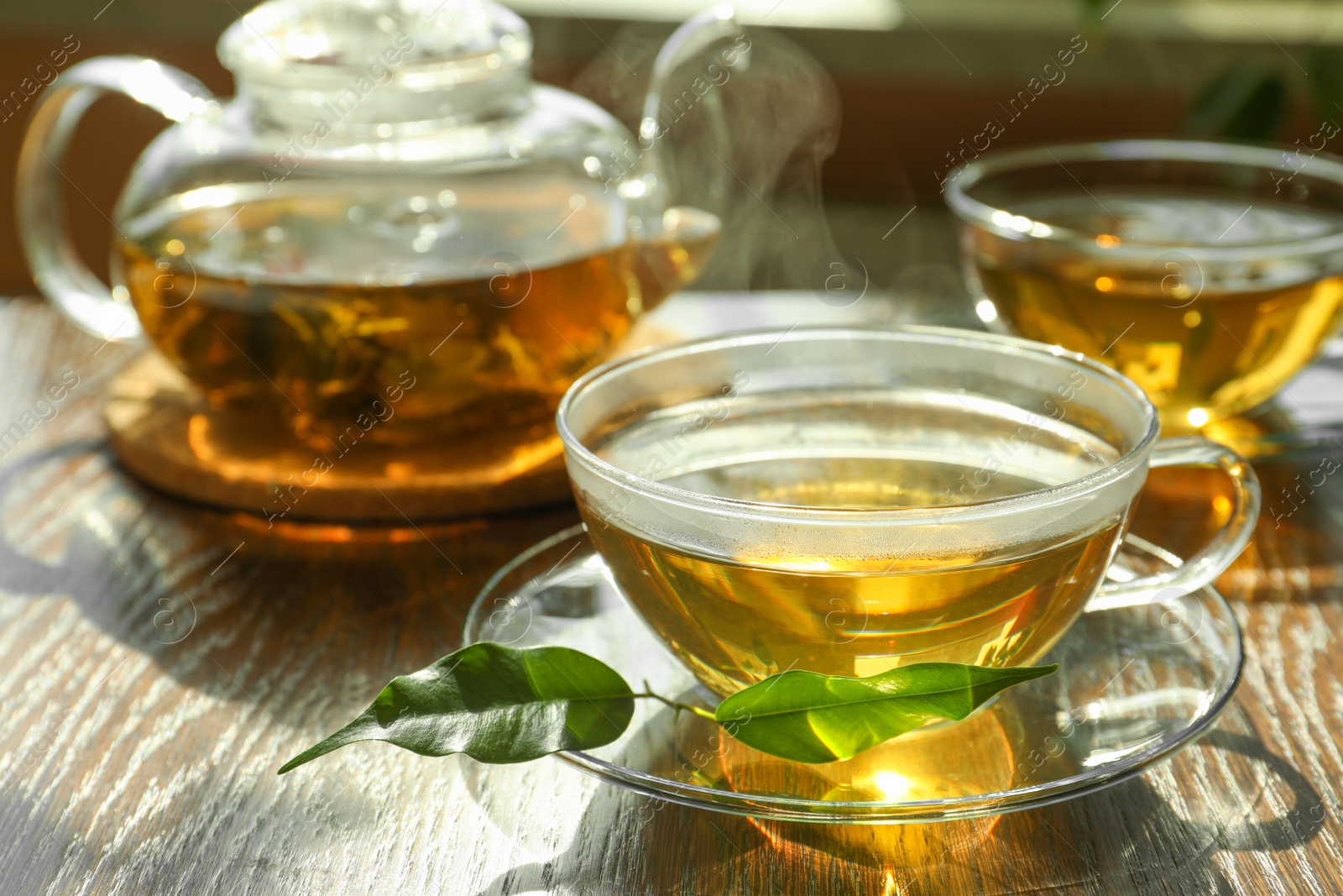 Photo of Fresh green tea in glass cups with saucers, leaves and teapot on wooden table