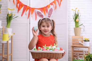 Photo of Adorable little girl with bunny ears and basket full of dyed eggs in Easter photo zone