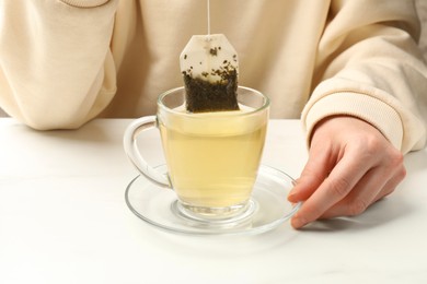 Photo of Woman putting tea bag in glass cup with hot water at white table, closeup