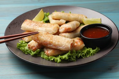 Photo of Plate with tasty fried spring rolls, lettuce, lime and sauce on light blue wooden table, closeup