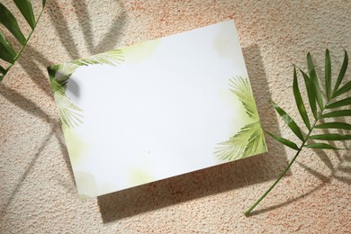 Blank invitation card and green leaves on light textured table, flat lay