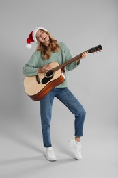 Photo of Young woman in Santa hat playing acoustic guitar on light grey background. Christmas music