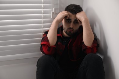 Sad young man near closed blinds at home