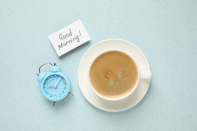 Photo of Delicious coffee, alarm clock and card with GOOD MORNING wish on light background, flat lay