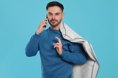 Man holding garment cover with clothes while talking on phone against light blue background. Dry-cleaning service