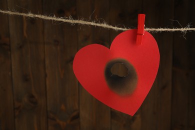 Photo of Red paper heart with burnt hole on rope near wooden wall, space for text. Broken heart