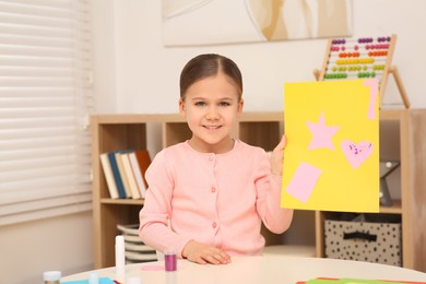 Cute little girl with colorful paper card at desk in room. Home workplace