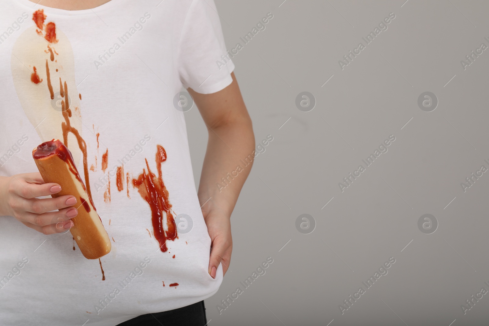 Photo of Woman holding hotdog and showing stain from sauce on her shirt against light grey background, closeup. Space for text