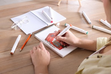 Photo of Woman drawing in sketchbook with felt tip pen at wooden table, closeup