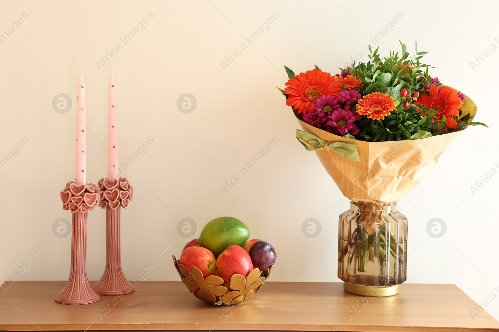 Photo of Bouquet of flowers, bowl with fresh fruits and candles on wooden table near white wall