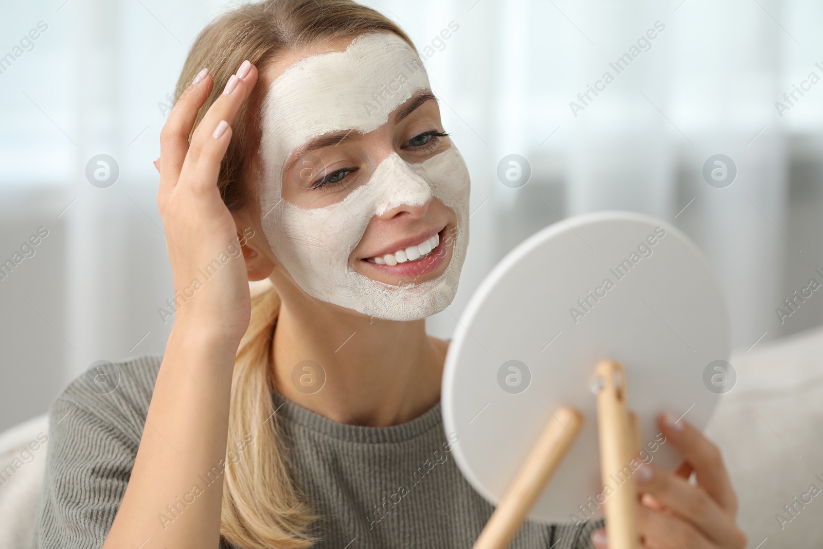 Photo of Young woman with face mask looking into mirror indoors. Spa treatments