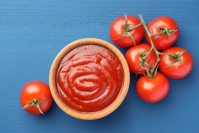 Photo of Bowl of tasty ketchup and tomatoes on blue wooden table, flat lay