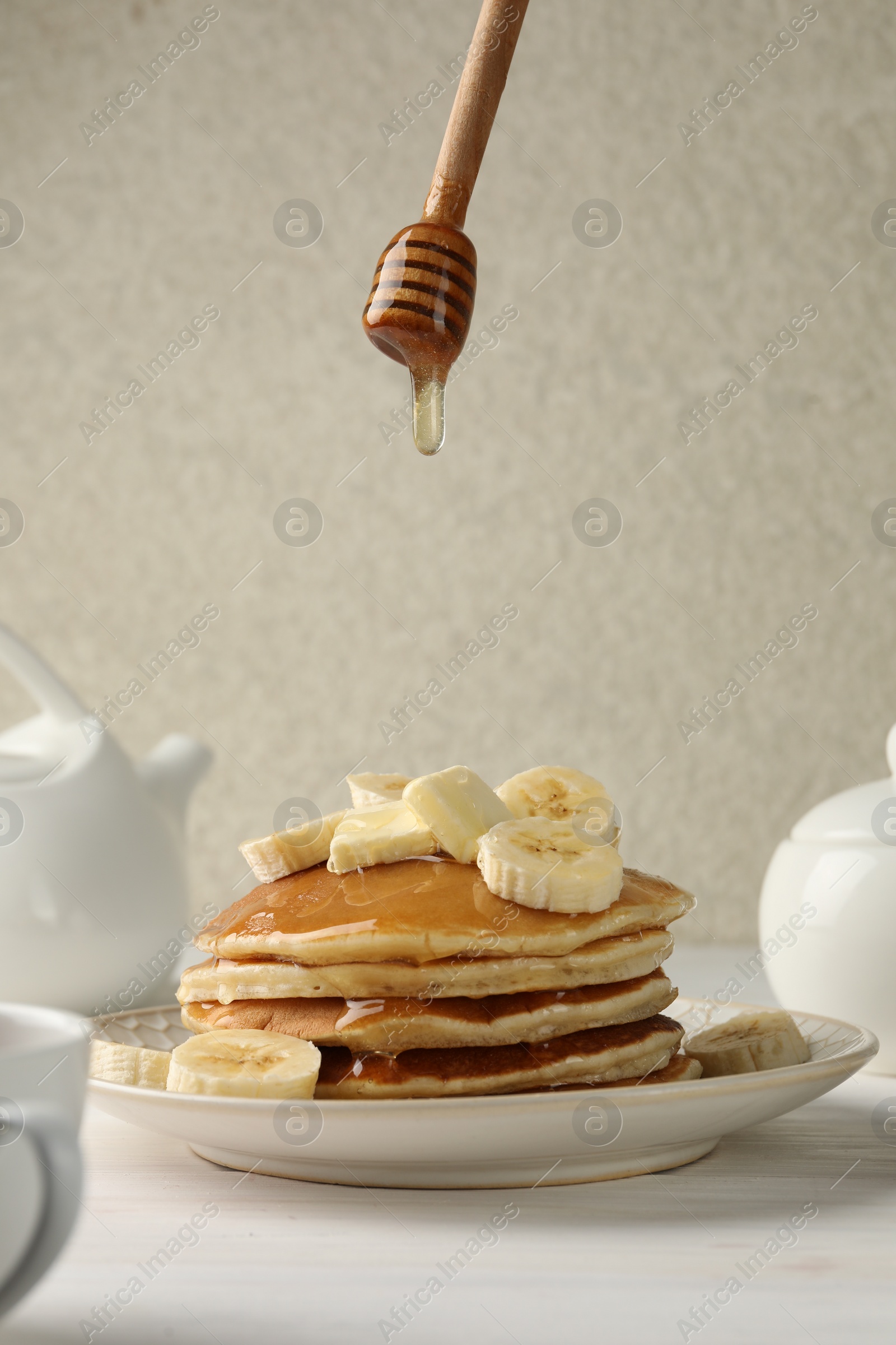 Photo of Dripping honey from dipper onto delicious pancakes with bananas and butter at white table