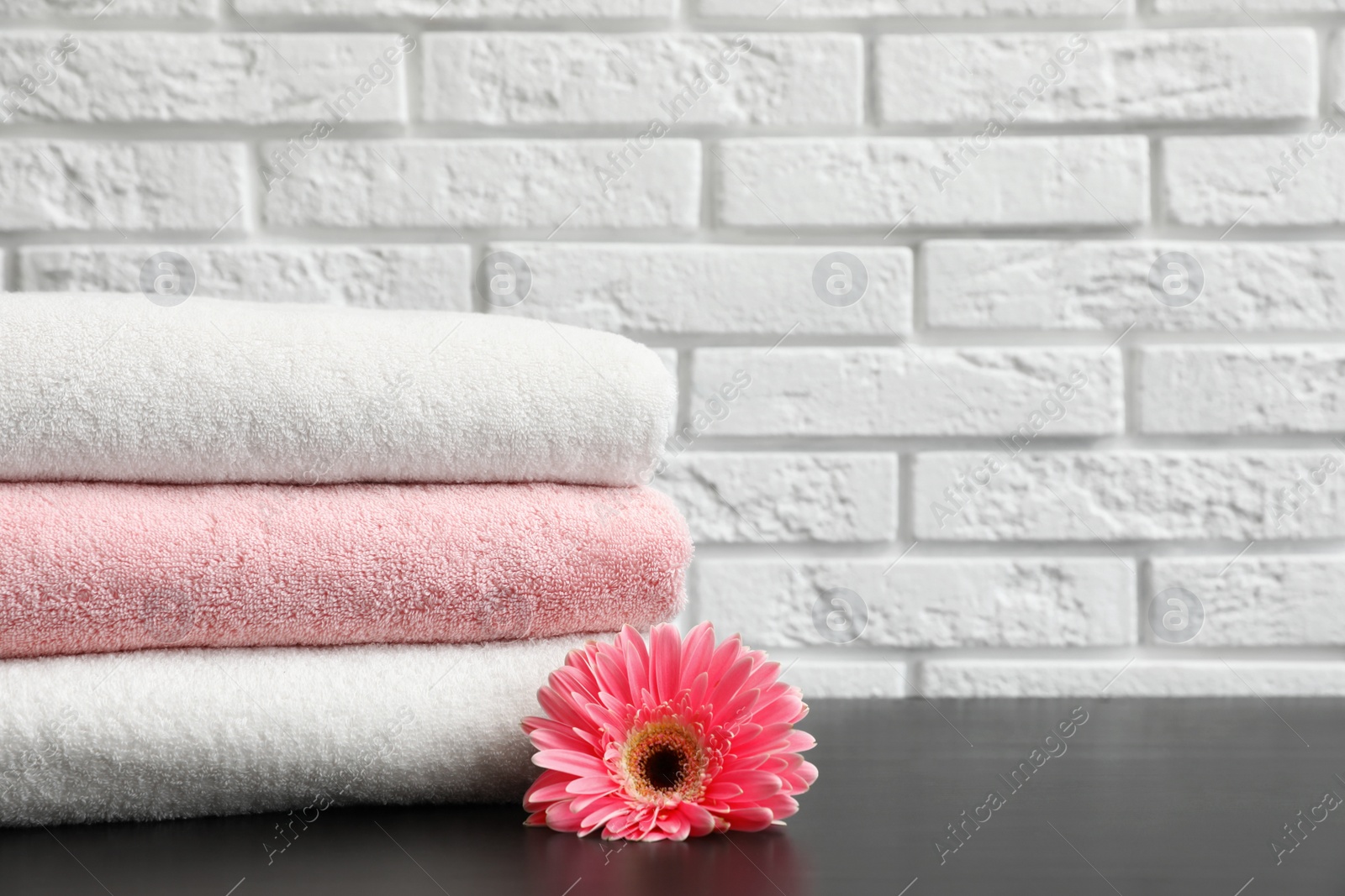 Photo of Stack of folded soft towels with flower on table near brick wall. Space for text