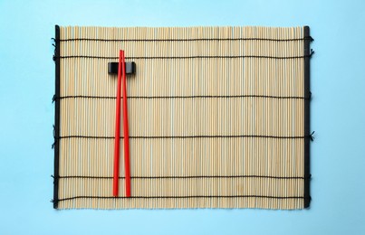 Photo of Bamboo mat with pair of red chopsticks and rest on light blue background, top view