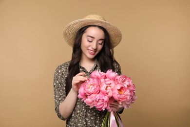 Photo of Beautiful young woman in straw hat with bouquet of pink peonies against light brown background