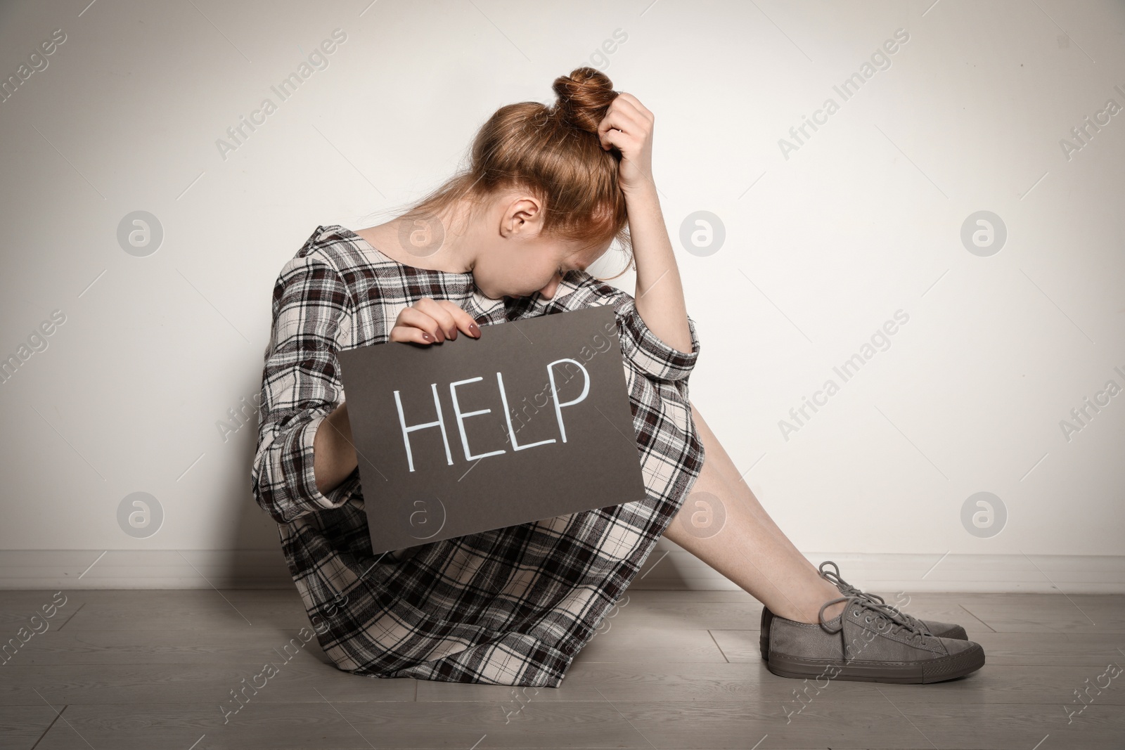 Photo of Young woman holding card with word HELP while sitting near light wall