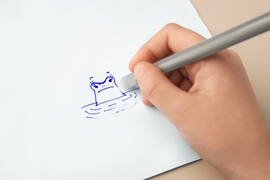 Child erasing drawing with erasable pen on paper sheet against beige background, closeup