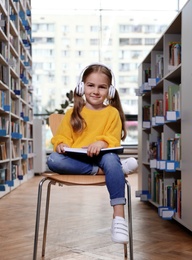 Photo of Cute little girl with headphones reading book on chair in library