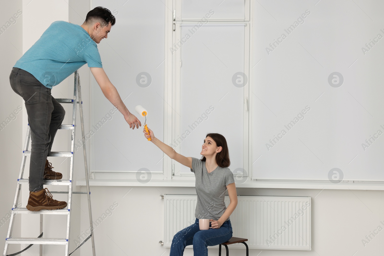 Photo of Young woman giving roller to man on stepladder indoors. Room renovation