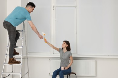 Photo of Young woman giving roller to man on stepladder indoors. Room renovation
