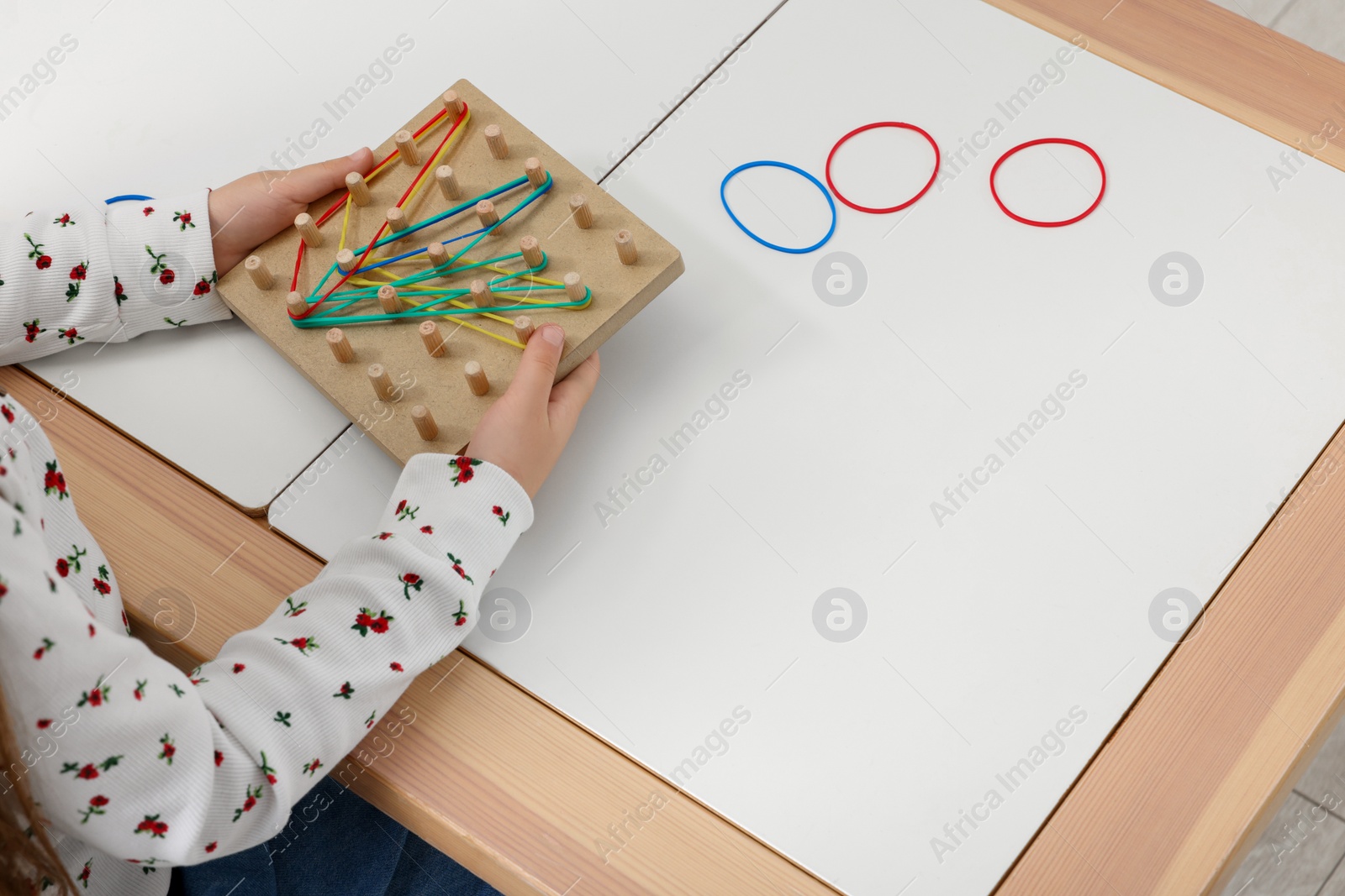 Photo of Motor skills development. Girl playing with geoboard and rubber bands at white table, closeup. Space for text