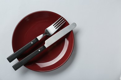 Photo of Burgundy ceramic plate with cutlery on light grey background, top view. Space for text