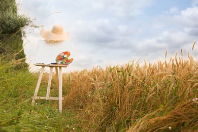 Wooden easel with blank canvas, painting equipment and hat in field. Space for text