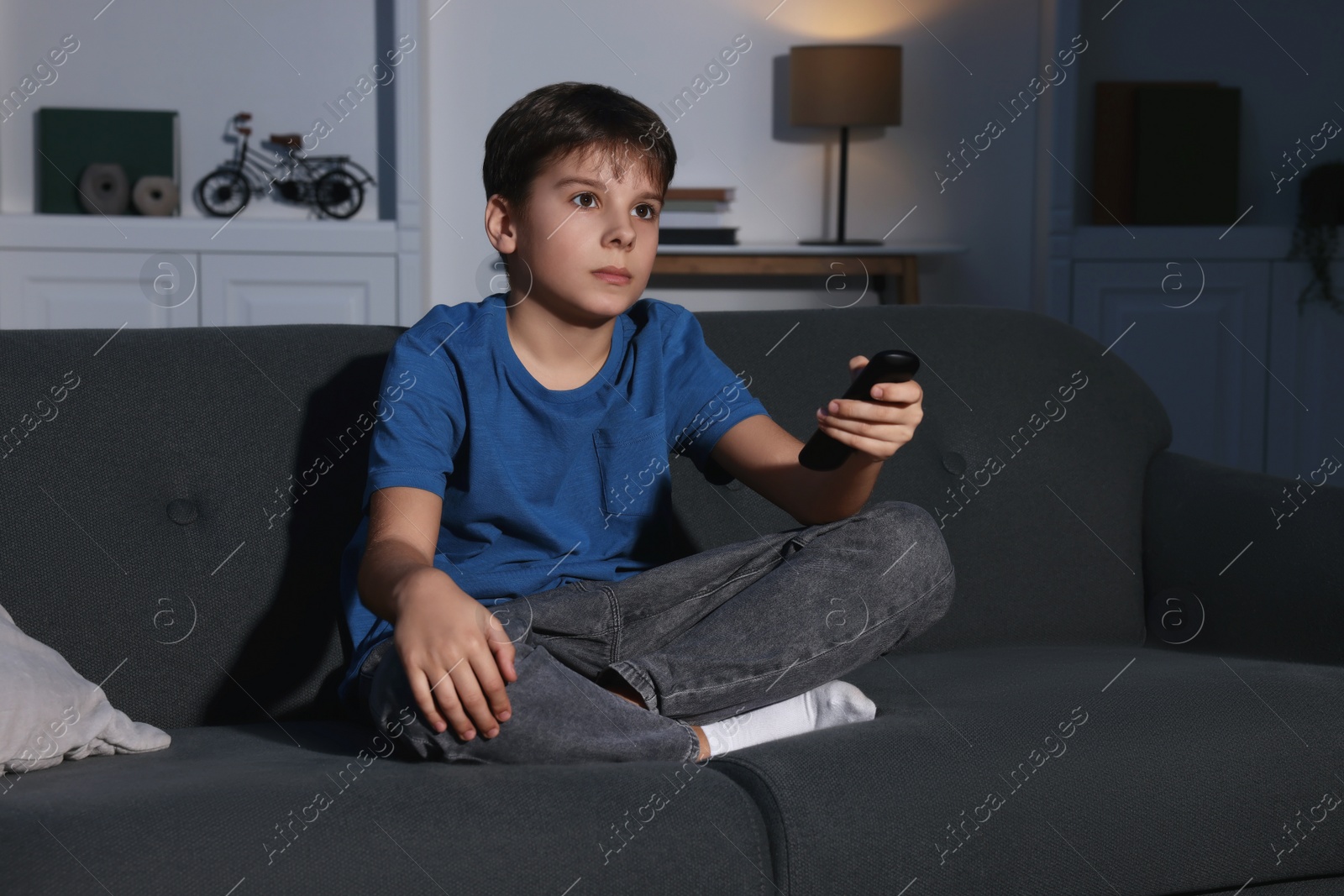 Photo of Little boy changing TV channels with remote control on sofa at home