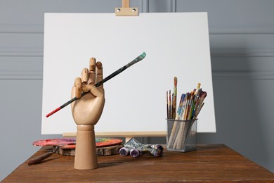 Easel with blank canvas, hand model and different art supplies on wooden table near grey wall