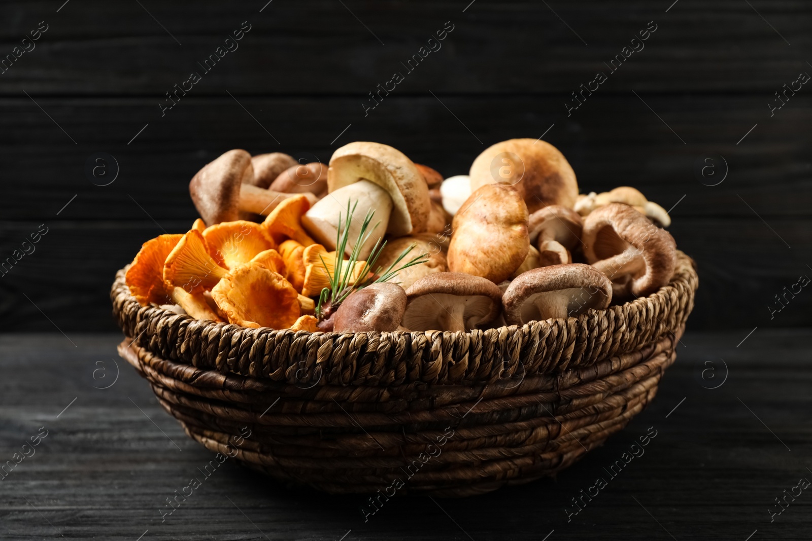 Photo of Different fresh wild mushrooms in wicker bowl on black wooden table, top view