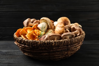 Photo of Different fresh wild mushrooms in wicker bowl on black wooden table, top view
