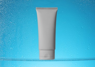 Tube with moisturizing cream on light blue background, view through wet glass