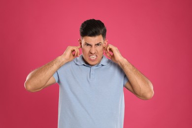 Emotional man covering ears with fingers on pink background