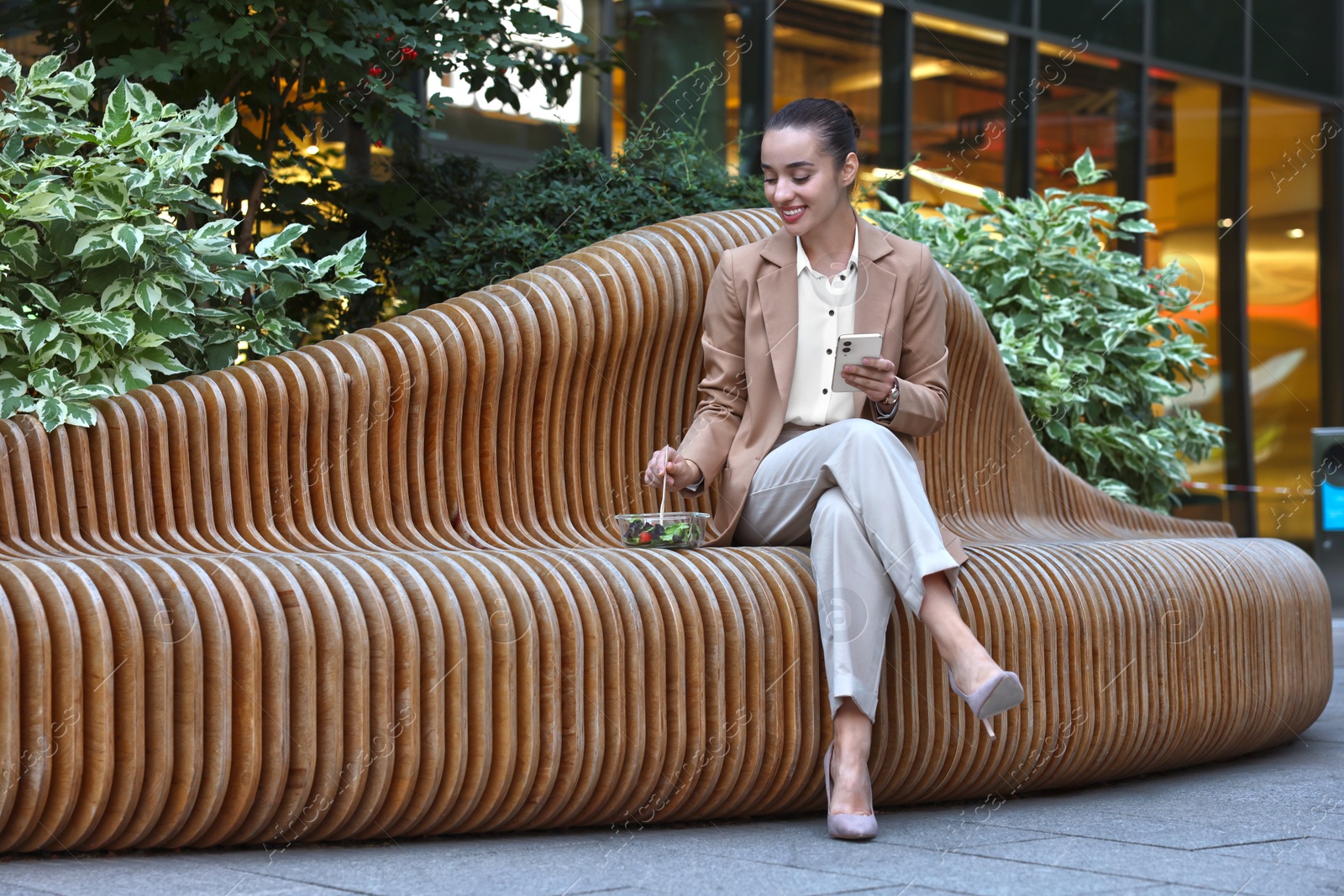 Photo of Happy young businesswoman with smartphone eating lunch on bench outdoors