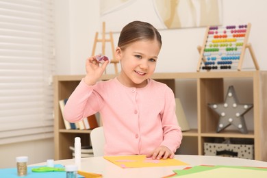 Photo of Cute little girl with container of colorful glitter at desk in room. Home workplace