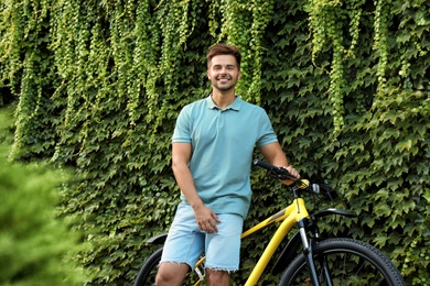 Photo of Handsome young man with bicycle near wall covered with green ivy vines on city street