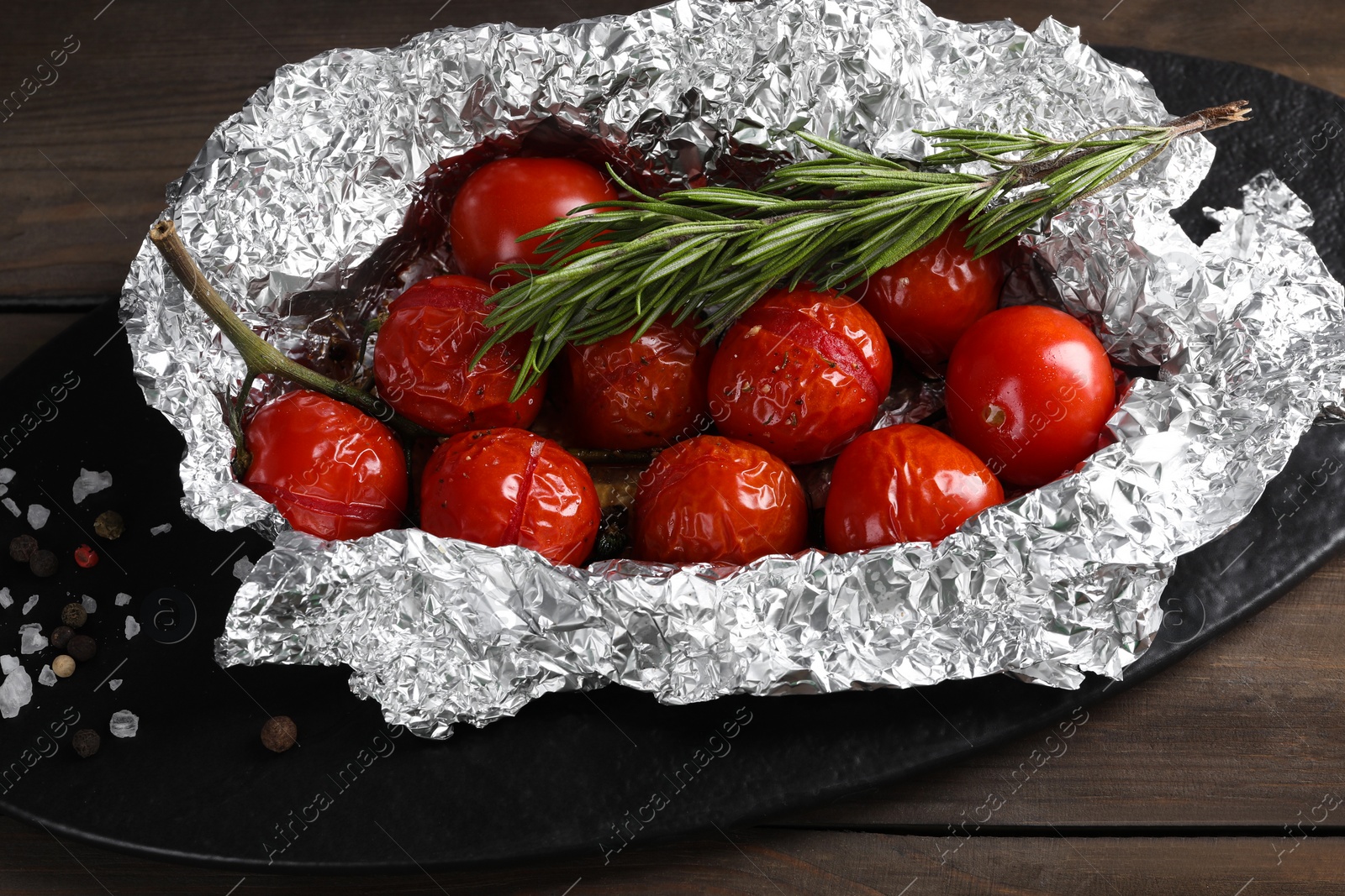 Photo of Aluminum foil with delicious baked tomatoes and rosemary on wooden table, closeup