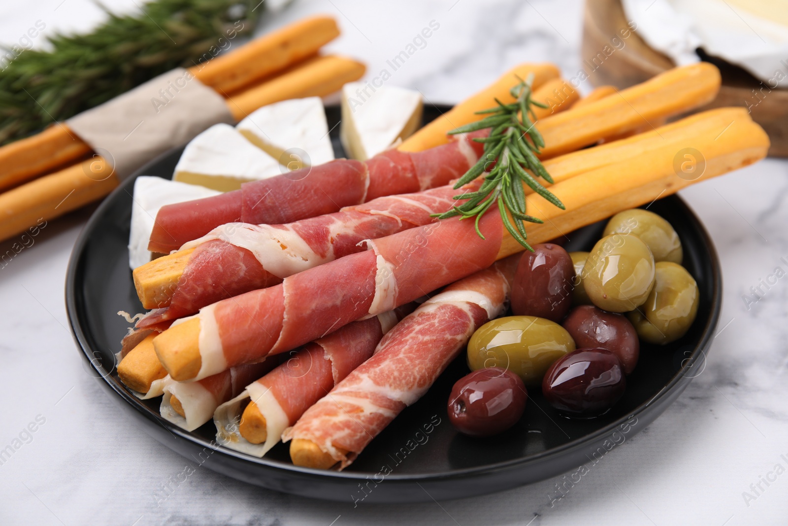 Photo of Delicious grissini sticks with prosciutto, cheese and olives on white marble table, closeup