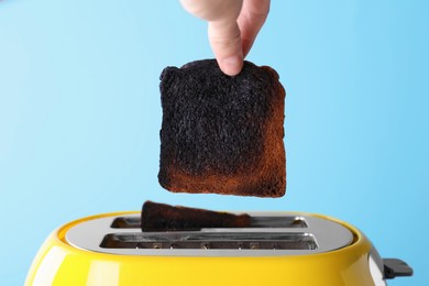 Photo of Woman taking off burnt bread from toaster against light blue background, closeup
