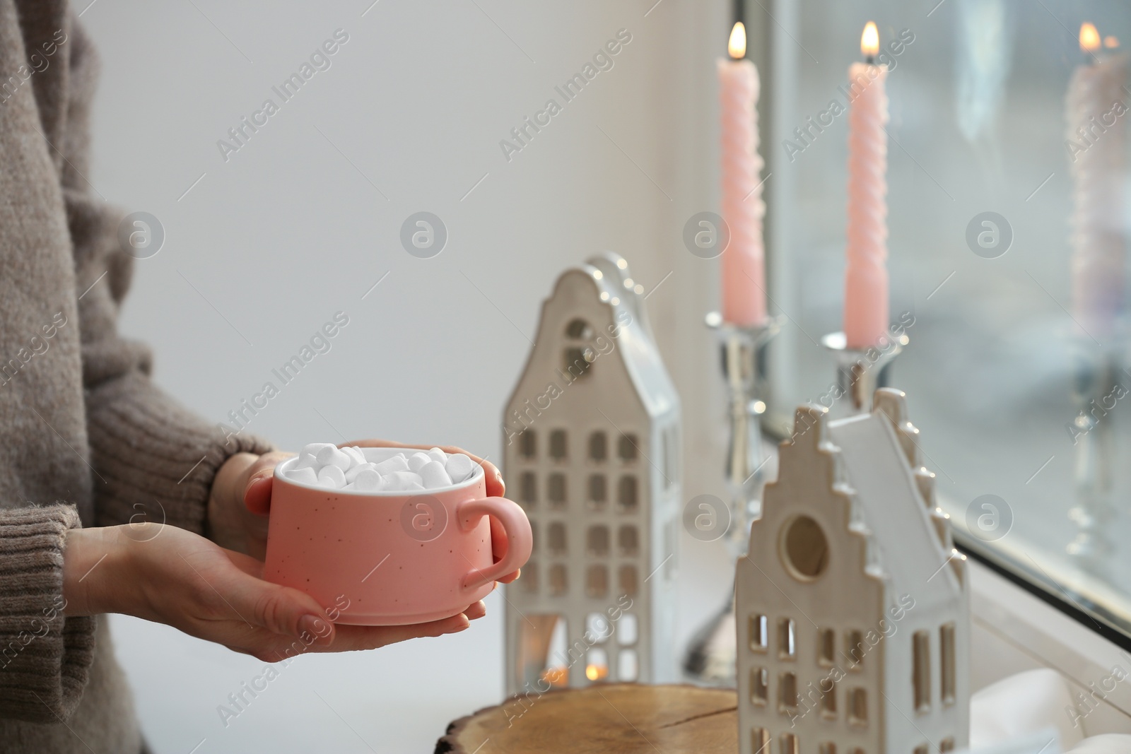 Photo of Woman holding cup of delicious drink near windowsill with house shaped lanterns indoors, closeup. Space for text